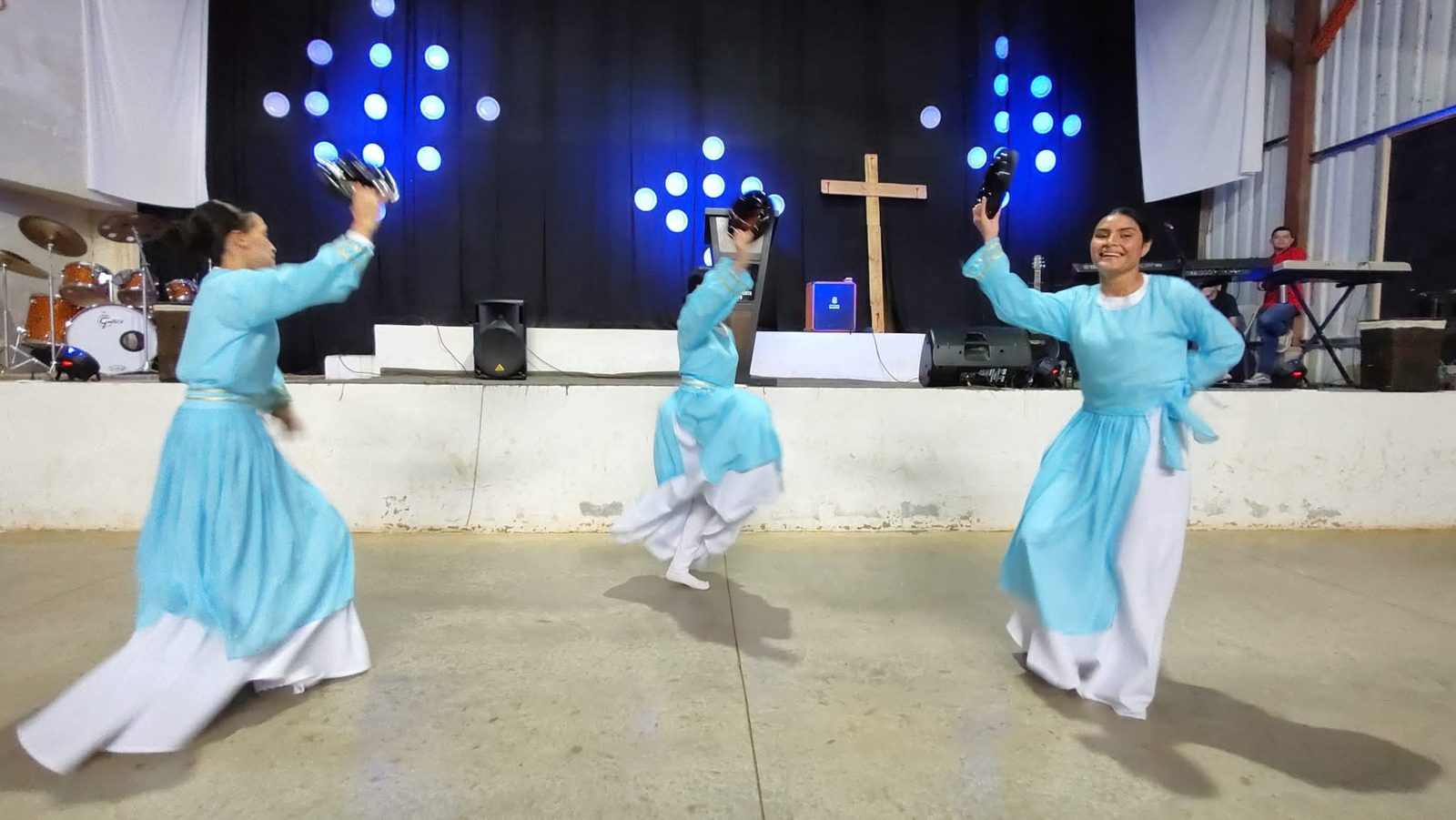 Danzarinas de Kemuel con vestuario turquesa y blanco danzan con panderos frente al escenario